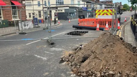 BBC/Richard Stead A large pile of dirt and rocks next to a big hole in a road which is surrounded by metal barricades.