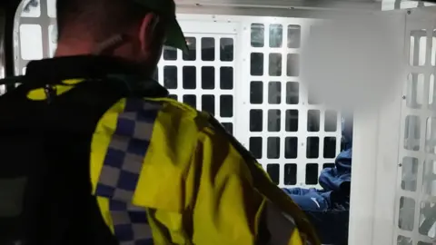 The back head and shoulders of a police officer is in the foreground on the left. In the background a suspect is sitting in a white barred enclosed cage in the back of a police van. His face is blurred.