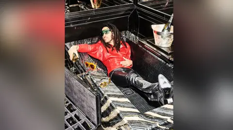 Tom Jackson Lenny is lounging in an an empty black tiled bathtub. He has a glass of champagne in one hand and a bottle in a silver ice bucket beside the bath. He is wearing a red leather jacket and black leather trousers. He has dreadlocks and sunglasses.