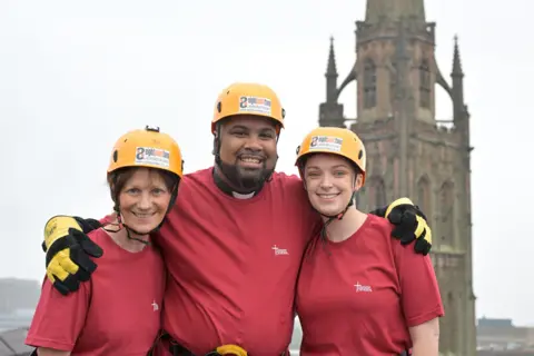 Coventry Cathedral Three volunteers wearing identical yellow hard hats and red t-shirts. A man in the middle wears a dog collar and is flanked by two women. They are all smiling at the camera with the tower of Coventry's old cathedral in the background. 