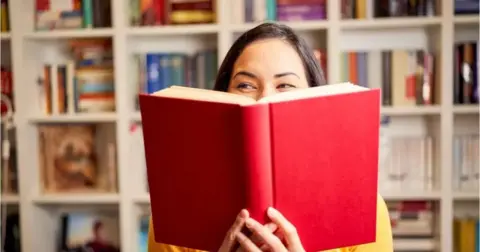 A woman holding up a red book which partially obscures her face - only her eyes and the top of her head can be seen. Behind her, and out of focus, are library bookshelves