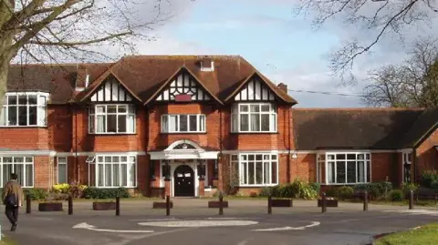 Geograph/David Hawgood A large, traditional brick building set back from a circular driveway. It features decorative timber framing on the upper sections and multiple white-framed windows, giving it a classic, institutional appearance. Leafless tree branches frame the scene, suggesting a cooler season, while small planters and bollards line the front entrance area. A person is visible walking near the building.