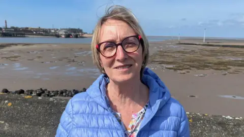 Karen Tunstall, local dog owner, wears a blue jacket and glasses and is standing in front of a beach.