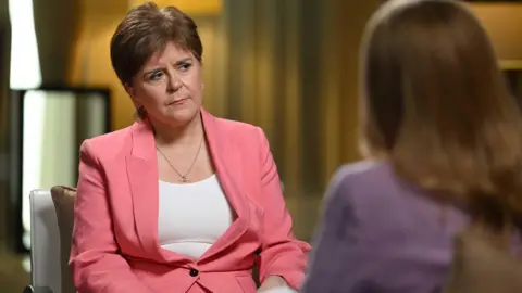 Nicola Sturgeon wearing a pink suit and white vest, with a quizzical expression. She is sitting on a chair facing another woman who has blonde hair and is wearing purple.