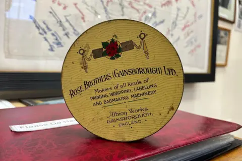 A round yellow tin stands on its side on a table in a museum. It features an image of a rose and the text: "Rose Brothers (Gainsborough) Ltd. Makers of all kinds of packing, wrapping, labelling and bagmaking machinery. Albion Works, Gainsborough, England." 
