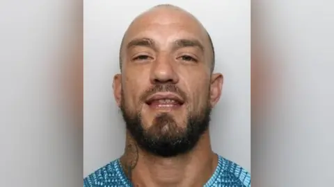 A mugshot of a man with a bald head and brown beard. He looks down his nose a slightly and holds his chin up, looking into the lens. He has a neck tattoo and wears a blue t shirt.