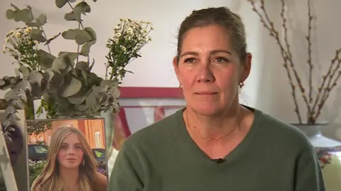 Sarah Renton sitting by a picture of her daughter. She is wearing a green jumper, has light brown hair, which is tied back, and gold star earrings. there are some dried flowers on display behind her.