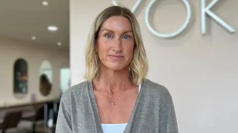 Jo Dyer is stood in front of plain white wall in her salon. She is staring straight at the camera. There is salon chairs and mirrors in the background slightly blurred. 