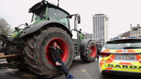 PA Media A man leans on a tractor tyre with a police car beside it that says GARDA