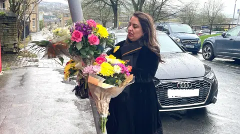 Charles Heslett/BBC The MP Naz Shah observes bunches of flowers stuck to a lamp-post on a public street. Some parked cars are behind her.