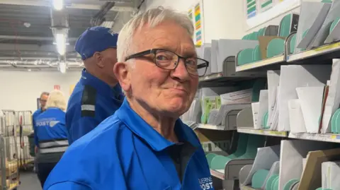 An old man with grey hair and glasses, wearing a blue polo shirt, smirks just to the left of the camera. He is standing in front of a shelf with letters on it, and other workers wearing blue guernsey post uniforms.
