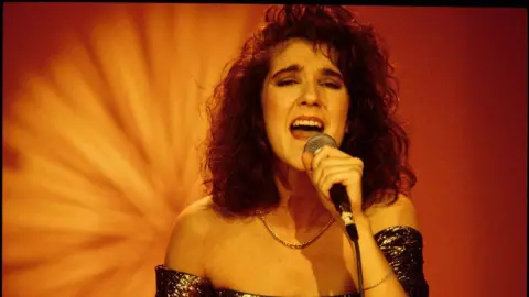 A young Celine Dion sings while holding a microphone. She is wearing a glittery strapless dress. 