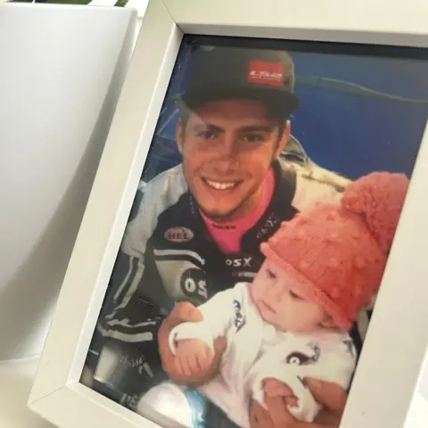 A picture of Daley Mathison wearing his motorbike leathers and a cap, holding Daisy Blu when she was a baby.