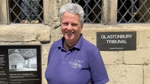 Wearing a purple polo shirt, Pauline is smiling at the camera, and standing next to a sign saying "Glastonbury Tribunal".