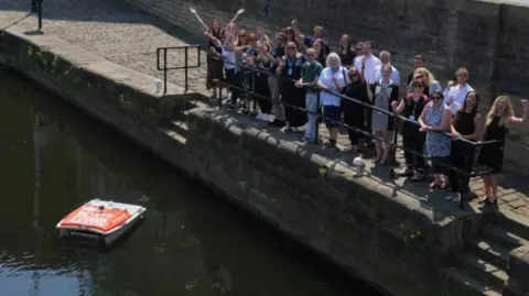 Leeds Waterfront Group A red robotic drone floating on the Leeds canal, with a crowd of 26 people standing on the dock side raising their arms and smiling.