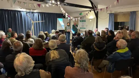 LDRS A community meeting in a hall where two presenters stand by a screen and a small wind‑turbine model, speaking to an audience seated beneath colourful bunting.