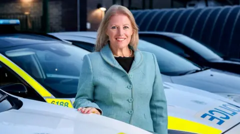 Hamphire and Isle of Wight PCC Donna Jones stands in a garage next to parked police cars. She has long, straight, fair hair and wears a blue jacket.
