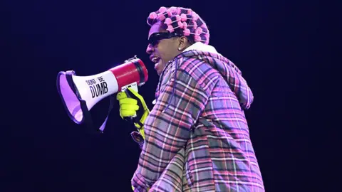 A$AP Rocky performing on stage. He wears a checked jacket and has pink rollers in his hair. He is holding a microphone with 'Don't Be Dumb' on it.