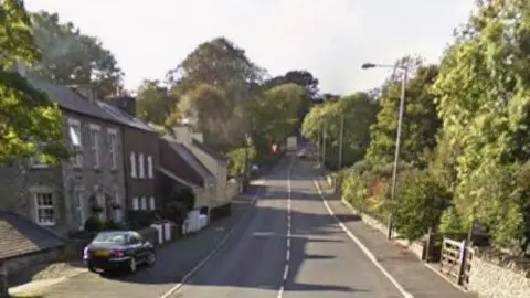 Google A view of the road, you can see a house to the left with a car parked outside, and a wall with a gate to the right and trees on bother side in the distance.
