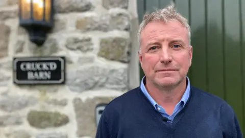 The picture shows Justin Heslop the director of a holiday home company called Peak Venues. He is wearing a blue jumper and is standing outside one of the properties