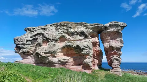 Ian Marshall Large weathered rock formation with layered red and grey tones, shaped into a natural arch on a grassy coastal cliff, with the sea visible beyond under a clear blue sky.