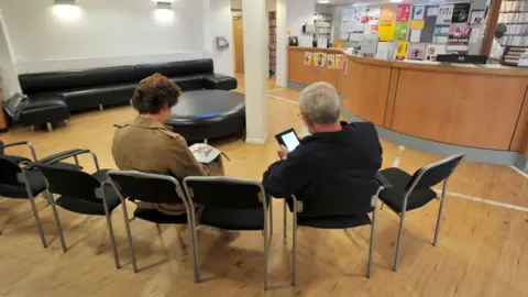 A man and woman are sitting with their backs to the camera on a row of chairs in a GP reception area