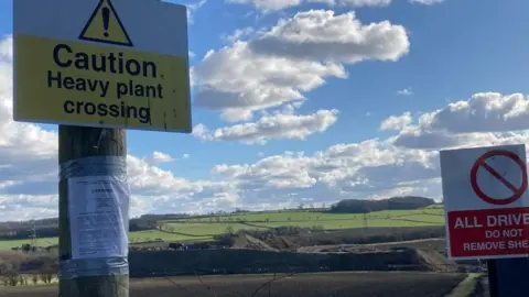 A yellow and white sign with black writing saying "Caution, heavy plant crossing" on a wooden pole that has an A4 piece of paper with writing stuck on with duct tape. Another sign is next to it with a red stop sign warning drivers. Fields are in the background as well as blue skies and some clouds. 