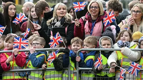 PA Media Primary school children in high vis vests waving flags behind a barrier. Behind them are smiling adults