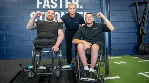 Two men in wheelchairs pose with their arms raised while another man stands behind them. They are in a fitness facility, with various pieces of gym equipment around in the background.