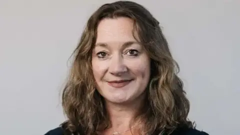 TYNWALD Daphne Caine, a woman with brown wavy hair smiles in front of a white backdrop.