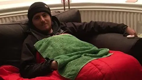 Myfanwy Sleep Tomos Sleep, wearing a hat and resting on sofa with a sleeping bag in 2023. He has a ginger-coloured beard and appears tired. The light is dimmed.