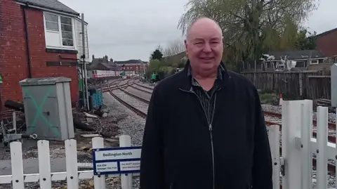 Dennis Fancett is standing at Bedlington Station with two of rail tracks behind him and a signal box to his right. He is a man in his 60s wearing a black jacket