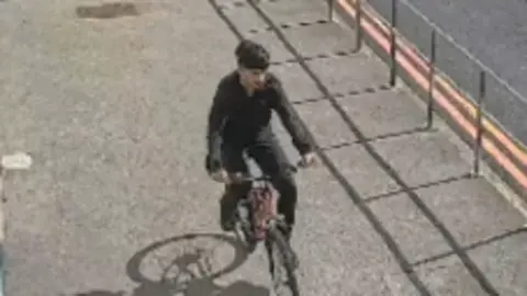 Lancashire Police CCTV overhead still of a young male with dark crop of hair in a black long-sleeved top and trousers riding a bike on a pavement next to a hospital road