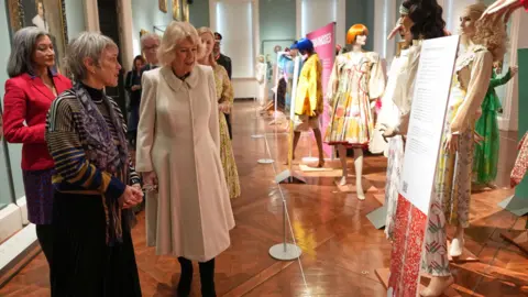 PA Media The Queen is walking past an exhibition of dresses on mannequins. The dresses are colourful. People are next to the Queen explaining the exhibition to her.