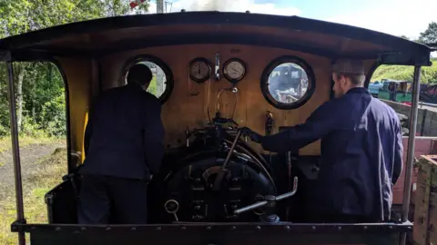 Tanfield Railway The footplate of a steam engine has two men standing on it who are driving the train. They are both wearing blue overalls and looking through the wooden portholes onto the track ahead 