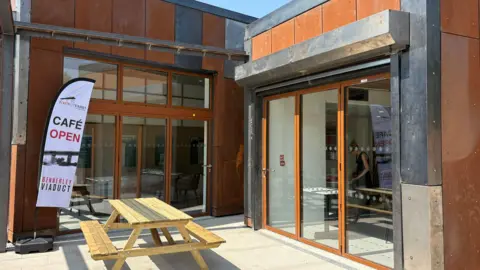 An orange steel building with glass doors and a flag which says Cafe Open