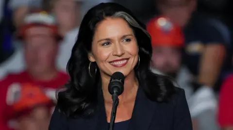 Reuters Former U.S. Rep. Tulsi Gabbard attends a campaign rally of Republican presidential nominee and former U.S. President Donald Trump at PPG Paints Arena in Pittsburgh, Pennsylvania - she is standing at the mic, has a broad smile, and gold earrings with a dark jacket and top. She has a distinctive white streak in her hair, and a blurred crowd can be seen behind her.