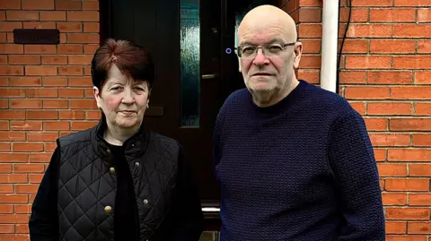Jayne has short reddish dark hair and is wearing a black gilet and jumper. Peter is a bald man with glasses and is wearing a blue jumper. They are standing in front of a red brick modern house and look serious. They are perhaps in their 50s.