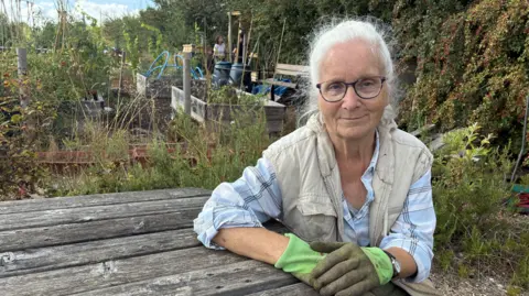 Philippa Slatter is sitting at a bench in an allotment. She has grey hair and is wearing glasses and gardening gloves.