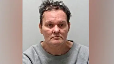 Police mugshot of Adam Saunders, who has brown unruly hair. He is photographed wearing a grey top.