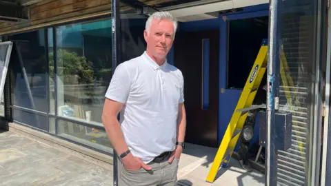 Scott Douglas looking straight at the camera. He is wearing a white polo shirt and is tucking his hands into the pockets of grey jeans. He is standing in front of a glass windowed office with a yellow ladder inside the door.