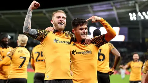 PA Media Hull City's Oliver McBurnie celebrates with Ryan Giles after scoring a goal at the MKM Stadium. McBurnie has blond hair and beard and Giles has black hair. The kit is amber shirts with black arm patterns and black shorts. Other Hull City players are in the background