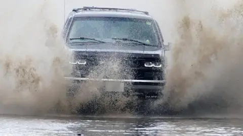 The picture shows a dark-coloured car driving straight through deep surface water, sending large walls of spray up on both sides of the vehicle. The water is being forced aside by the front of the car, creating a dramatic splash that almost reaches the height of the bonnet and partially obscures the headlights.
The road surface is completely covered by water, suggesting local flooding rather than a small puddle. 