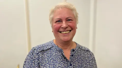 A woman with light blonde hair smiling widely towards the camera. She is wearing a blue flowery blouse. 