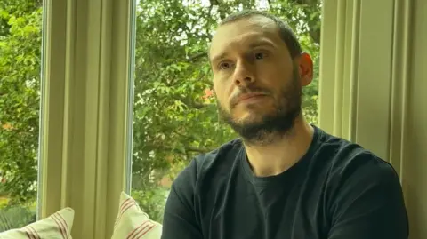 Joel Housego has short black hair and a beard and moustache. He is sitting indoors near a bay window and wears a black T-shirt. Outside there are trees in full leaf. There are two cushions behind him, primarily lime green with vertical red and white stripes. The walls and window frame are similarly lime green.