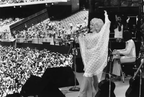Richard McCaffrey/Michael Ochs Archives/Getty Images Parton performs on stage in 1978, in a pale shawl and before a big crowd. 