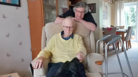 Diana Gibbs Mick sits in an armchair stroking his black labrador dog with his walking aid beside him. His wife Diana stands behind him smiling.