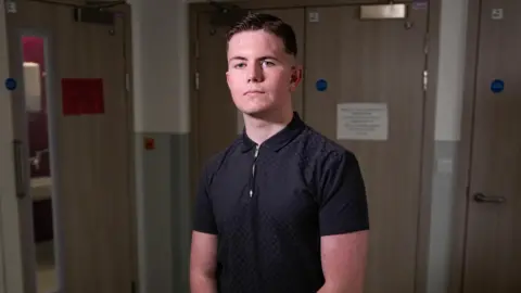 PA Media A young man looks at the camera - he has brown hair and is wearing a navy blue short-sleeve shirt. He appears to be in a hospital, with several wooden doors behind him