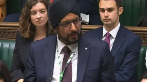 Mr Juss is stood up speaking in Parliament. He wears a turban and glasses and is dressed in a navy suit.