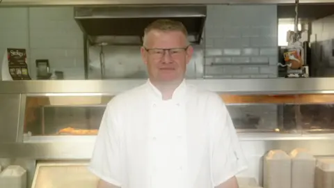 Andrew Crook stands in front of a fish shop counter, wearing chef's whites. A display counter and white tiled walls can be seen behind him. He has short grey hair and glasses.